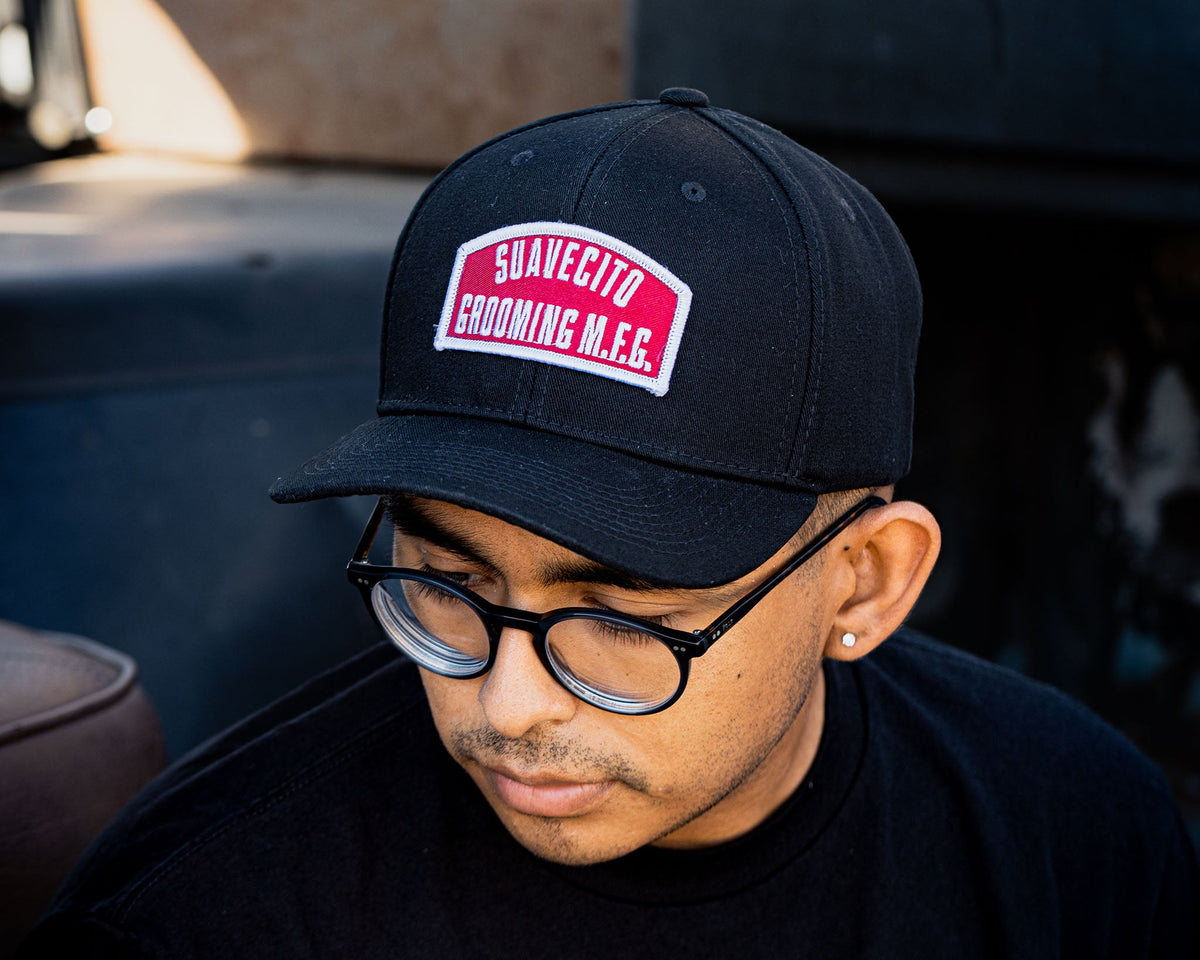 person wearing - suavecito hat with embroidered text and red background "suavecito grooming m.f.g."